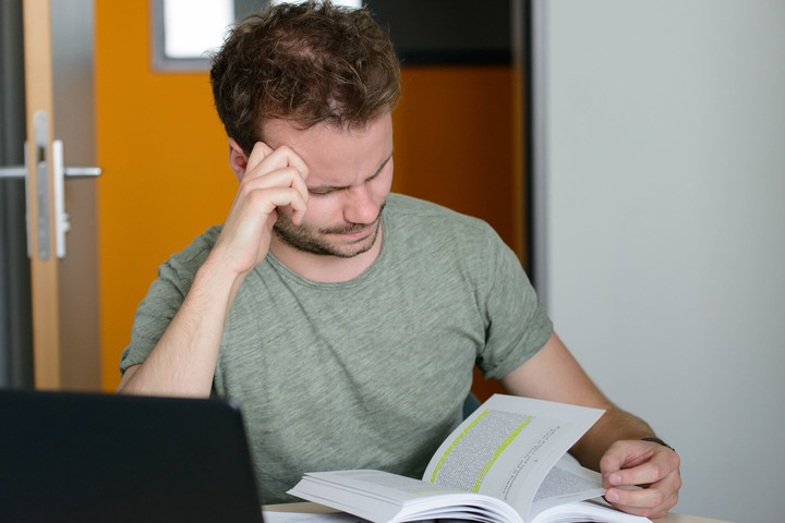 Student sitzt am Schreibtisch mit einem Laptop. Er blättert in einem Buch, hat seinen Kopf auf die Hand gestützt und schaut etwas verzweifelt. 
