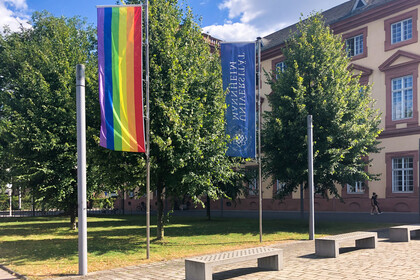 Regenbogenflagge und Flagge der Universität Mannheim wehen vor dem Ostflügel des Mannheimer Schlosses, Bäumen im Hintergrund.