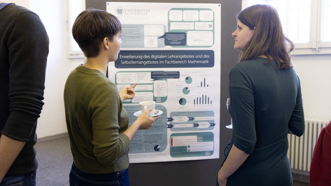 Zwei Frauen stehen vor einer Poster-Stellwand und unterhalten sich. Das Poster hat den Titel "Erweiterung des digitalen Lehrangebots und des Selbstlernangebotes im Fachbereich Mathematik".