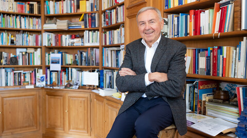 Eine lächelnde schick gekleidete Person sitzt auf einem Hocker vor einem großen Bücherregal. Die Person heißt Peter Eichhorn.