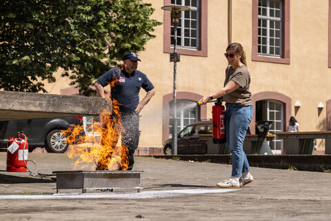 Eine Frau in Jeans löscht mithilfe eines Feuerlöschers ein kleines Feuer im Schneckenhof. Im Hintergrund ein Mann in dunkelblauer Arbeitskleidung.
