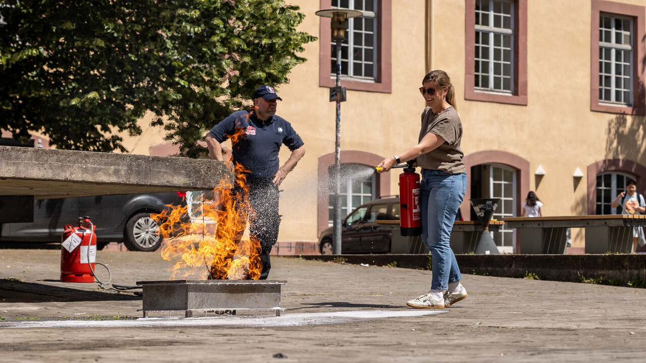 Eine Frau in Jeans löscht mithilfe eines Feuerlöschers ein kleines Feuer im Schneckenhof. Im Hintergrund ein Mann in dunkelblauer Arbeitskleidung.