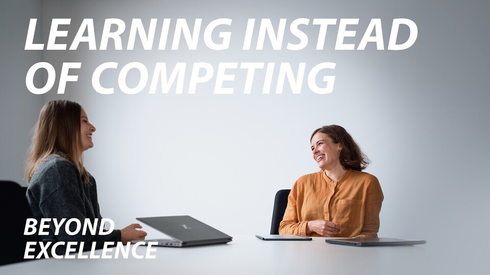 Two women sit across from each other at a white table, laughing, with two closed notebooks and a tablet. One wears an orange‑mustard blouse, the other a dark grey sweater. Text: “LEARNING INSTEAD OF COMPETING”; “Beyond excellence” (lower left).