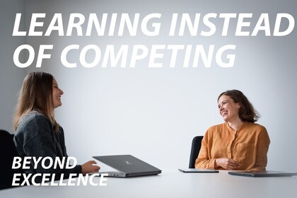 Two women sit across from each other at a white table, laughing, with two closed notebooks and a tablet. One wears an orange‑mustard blouse, the other a dark grey sweater. Text: “LEARNING INSTEAD OF COMPETING”; “Beyond excellence” (lower left).