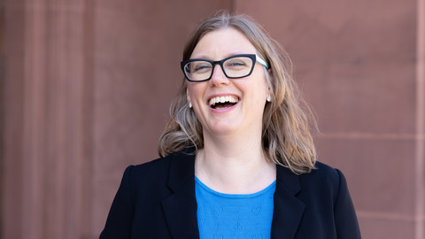 Eine lachende Person trägt eine blaue Bluse sowie einen schwarzen Blazer und steht vor einem braunen Gebäude. Die Person heißt Florence Oloff.