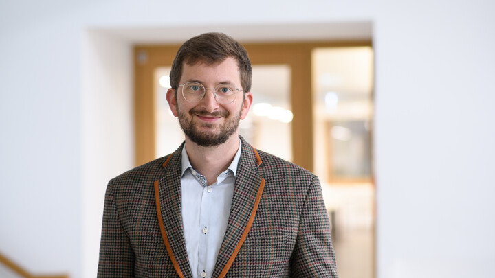 Philipp Müller steht in einem braun gemusterten Anzug vor einer Tür in einem Gebäude.