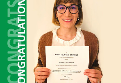 Elisa Casi-Eberhard. She is wearing a brown jacket over a white shirt and is holding a paper in front of a yellowish background.