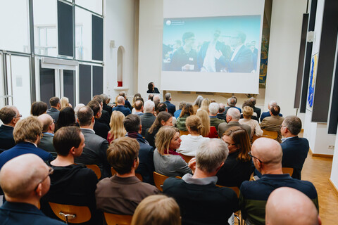 Das Bild zeigt einen vollbesetzten Festsaal aus der letzten Reihe. Im Hintergrund sieht man die Festrednerin am Rednerpult. Sie hat ihren Kopf zu einer Leinwand mit einer Präsentation gewendet. Die Leinwand zeigt fröhlich feiernde Menschen.
