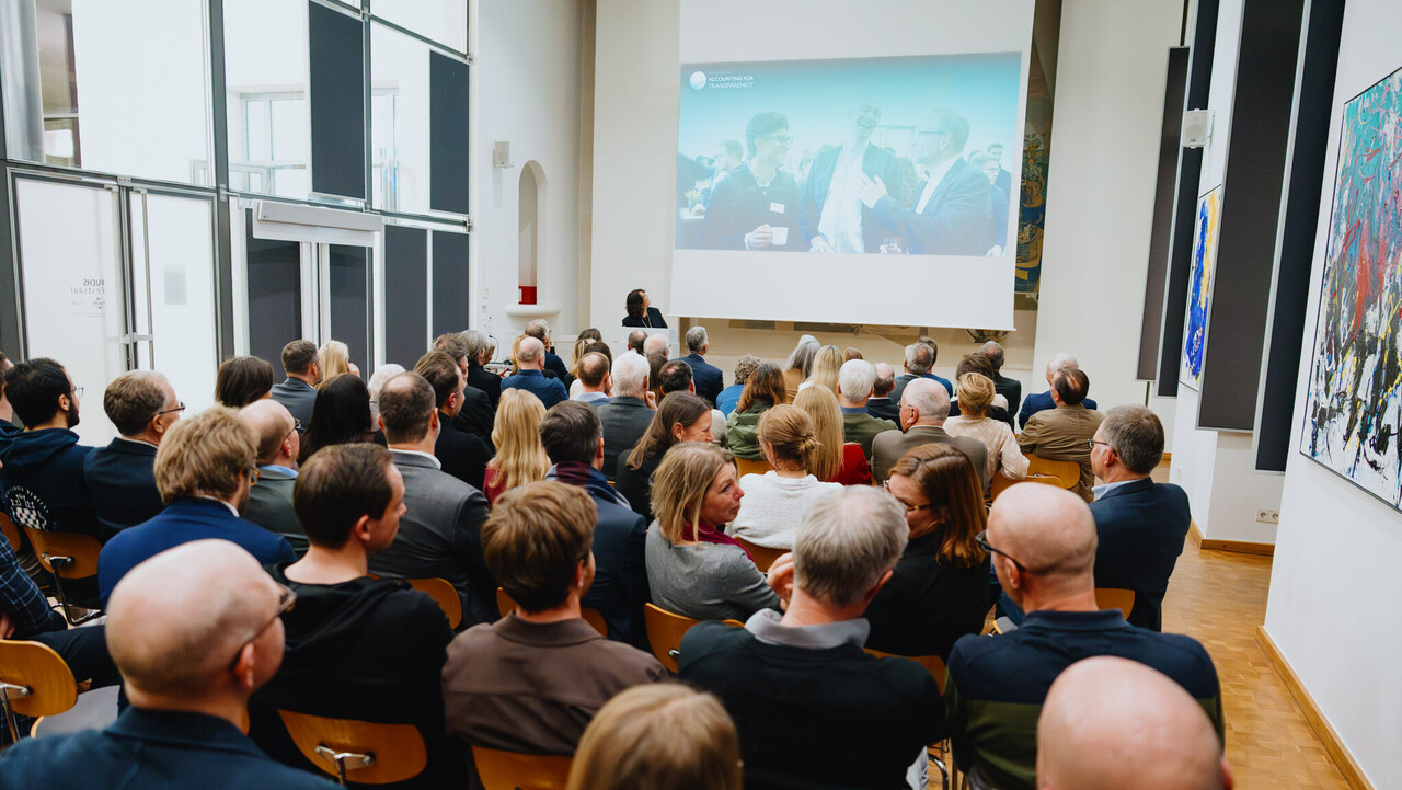 Das Bild zeigt einen vollbesetzten Festsaal aus der letzten Reihe. Im Hintergrund sieht man die Festrednerin am Rednerpult. Sie hat ihren Kopf zu einer Leinwand mit einer Präsentation gewendet. Die Leinwand zeigt fröhlich feiernde Menschen.