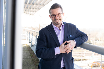 Eine lächelnde Person trägt ein lilafarbenes Hemd sowie einen dunkelblauen Blazer und lehnt an ein Geländer. Die Person heißt Nicolas Ziebarth.