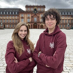 Eva Peetz and Amon Brehm stand laughing in front of Mannheim Palace. They are both wearing a wine-red hoodie and have their arms crossed.