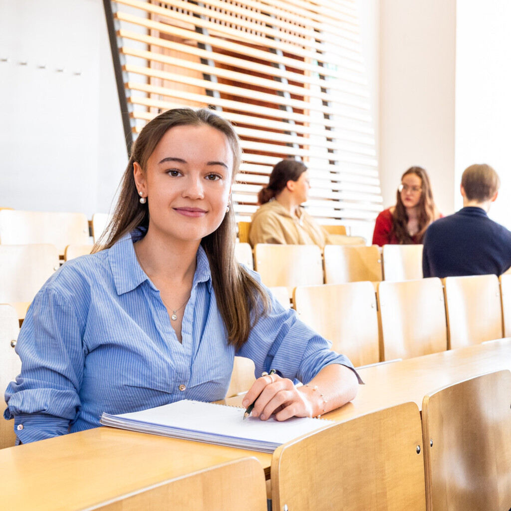 Studierende sitzen in einem Hörsaal. Sie schauen direkt in die Kamera.