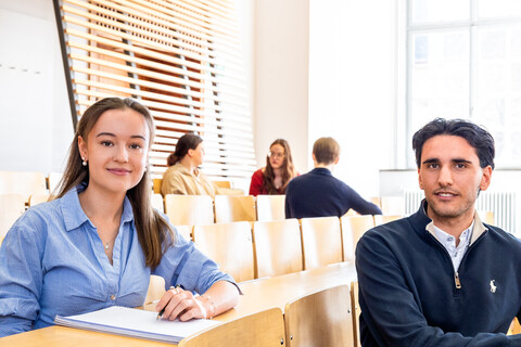 Studierende sitzen in einem Hörsaal. Sie schauen direkt in die Kamera.