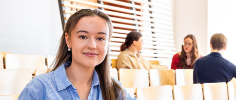 Studierende sitzen in einem Hörsaal. Sie schauen direkt in die Kamera.