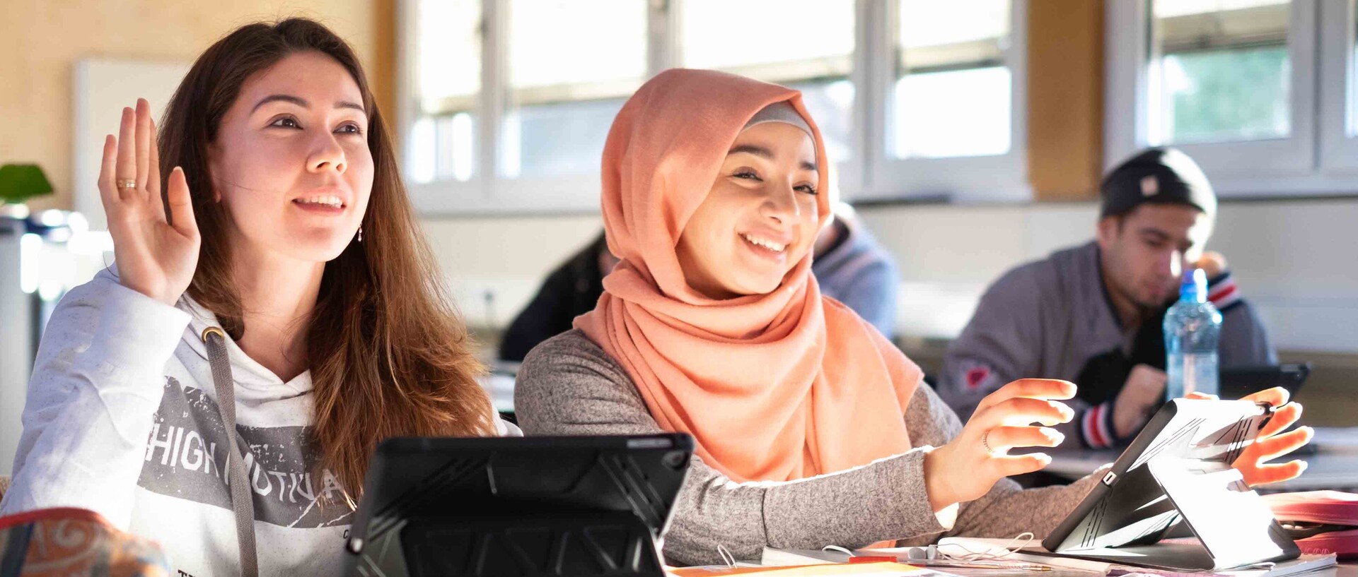 Zwei Schülerinnen sitzen in einem Klassenzimmer.  Eine hat lange offene Haare, die andere trägt ein apricotfarbenes Kopftuch.