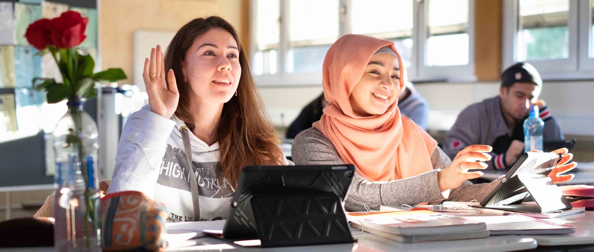 Zwei Schülerinnen sitzen in einem Klassenzimmer.  Eine hat lange offene Haare, die andere trägt ein apricotfarbenes Kopftuch. 