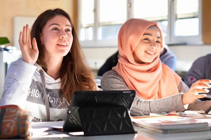 Zwei Schülerinnen sitzen in einem Klassenzimmer.  Eine hat lange offene Haare, die andere trägt ein apricotfarbenes Kopftuch. 