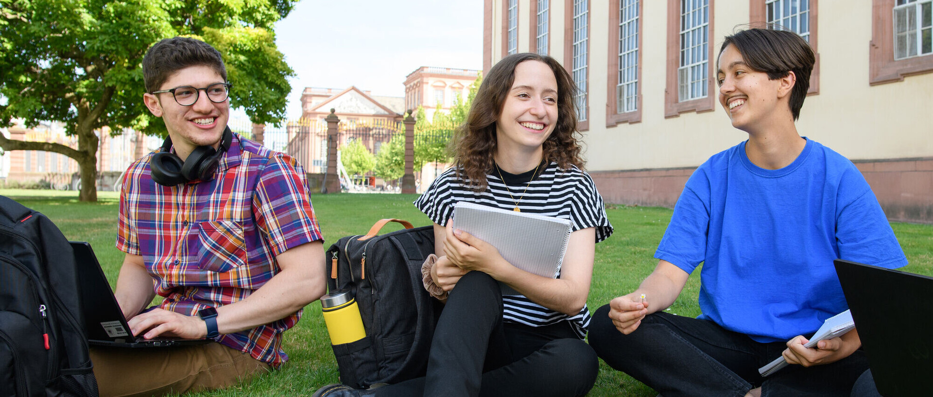 Lachende Studierende sitzen im Schneidersitz im Gras vor dem Schloss