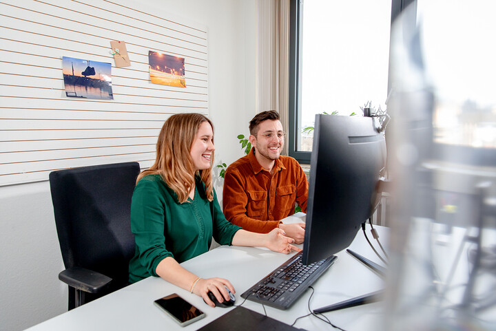 Zwei Mitarbeitende der Uni Mannheim sitzen lachend vor einem Bildschirm. 
