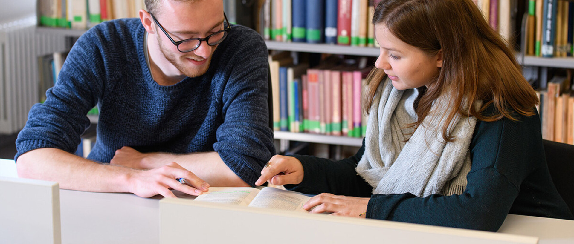 Eine Studentin zeigt einem Studenten etwas in einem aufgeschlagenen Buch.