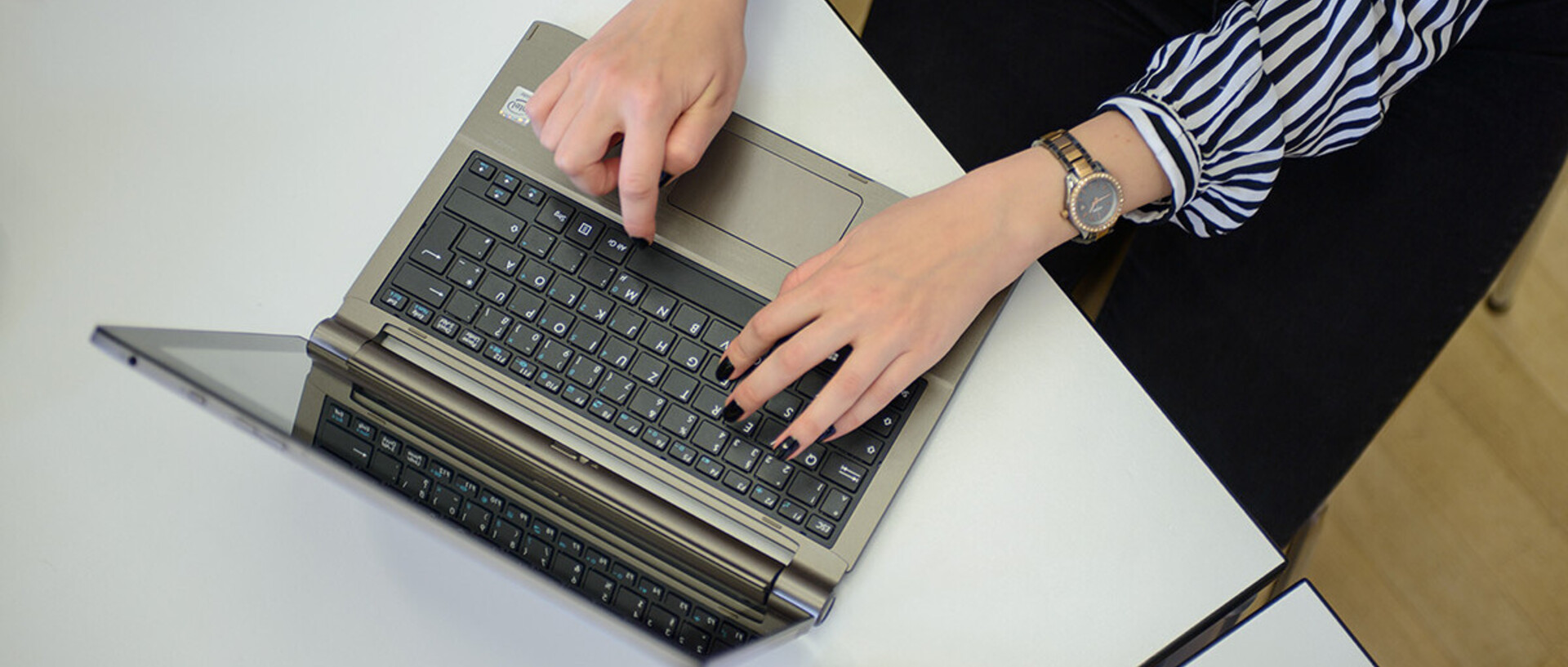 In einem Büro arbeitende Frau. Sie trägt eine schwarz weiß gestreifte Bluse, eine schwarze Hose und eine Uhr. Sie tippt etwas an einem Laptop.