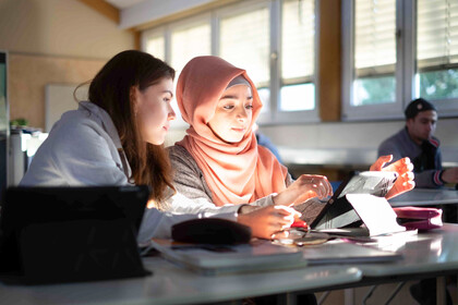 zwei Studentinnen sitzen am Tisch und schauen auf ein Tablet. 