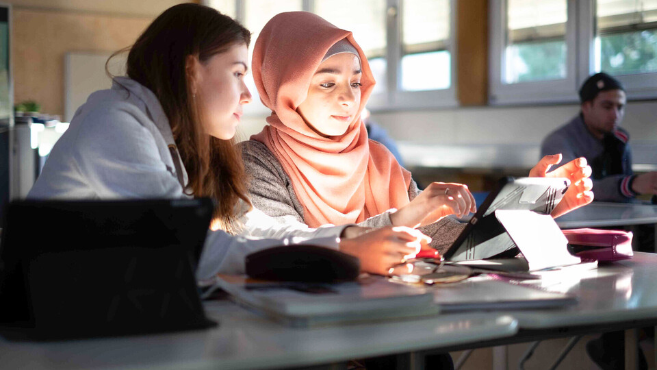 zwei Studentinnen sitzen am Tisch und schauen auf ein Tablet.