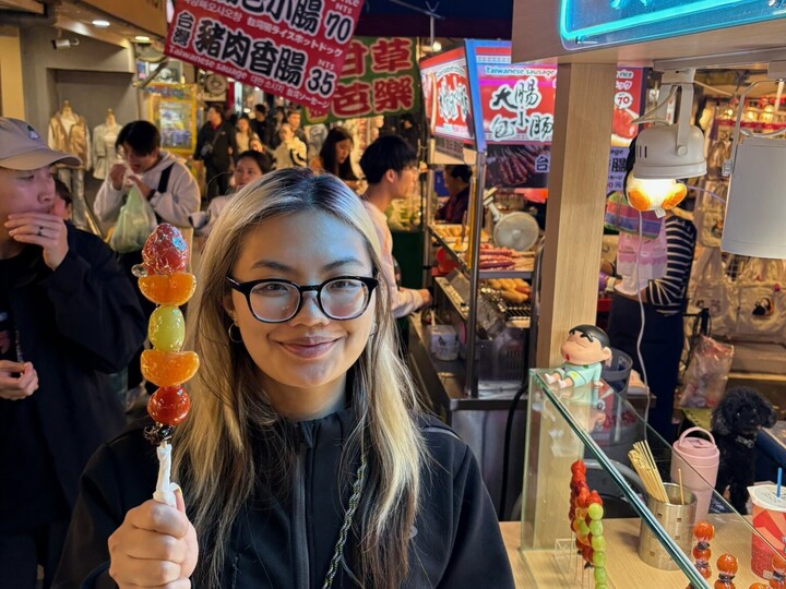Eine junge Frau mit blond gefärbten Haaren steht inmitten einer asiatischen Stadt, hält einen Früchtespieß in der Hand und lächelt in die Kamera..