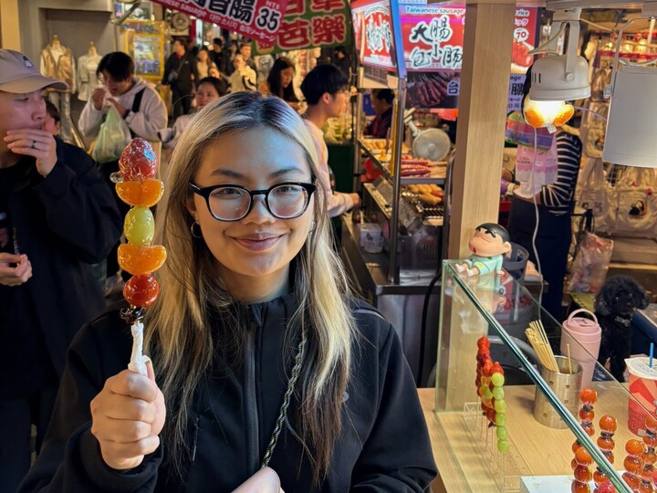 Eine junge Frau mit blond gefärbten Haaren steht inmitten einer asiatischen Stadt, hält einen Früchtespieß in der Hand und lächelt in die Kamera..