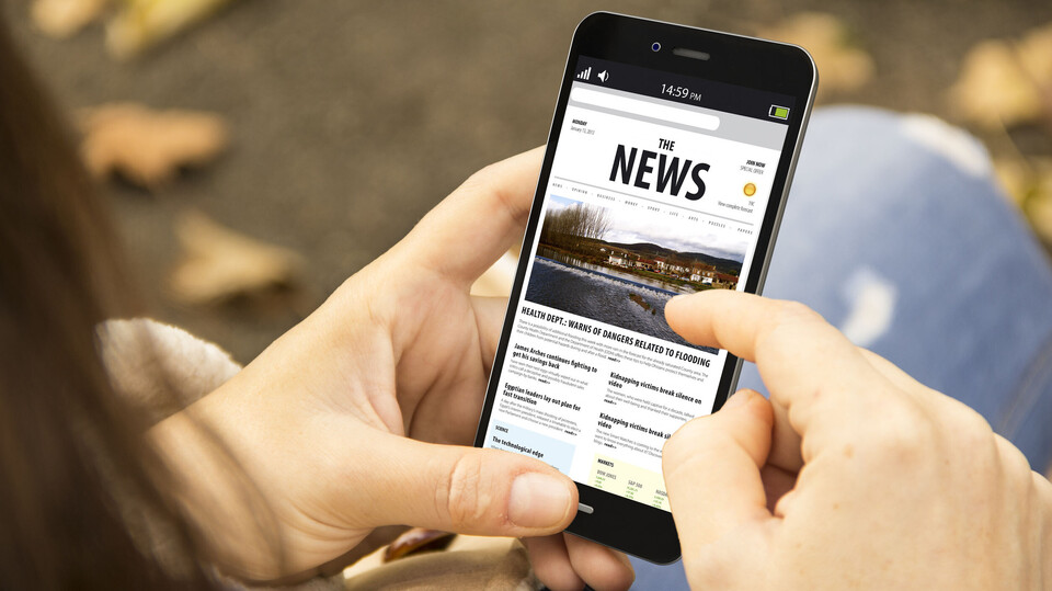 Eine Person hält ein Smartphone in der Hand. Auf dem Bildschirm ist das Wort "News" lesbar.