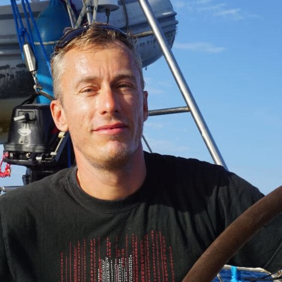 Stefan Kluge im Sonnenschein auf einem Segelboot. Er trägt ein schwarzes T-Shirt und eine Sonnenrille auf dem Kopf.