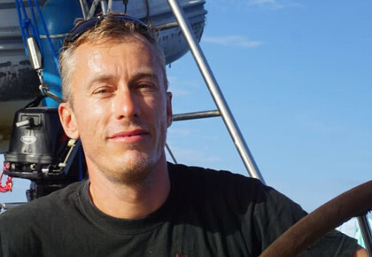 Stefan Kluge im Sonnenschein auf einem Segelboot. Er trägt ein schwarzes T-Shirt und eine Sonnenrille auf dem Kopf.