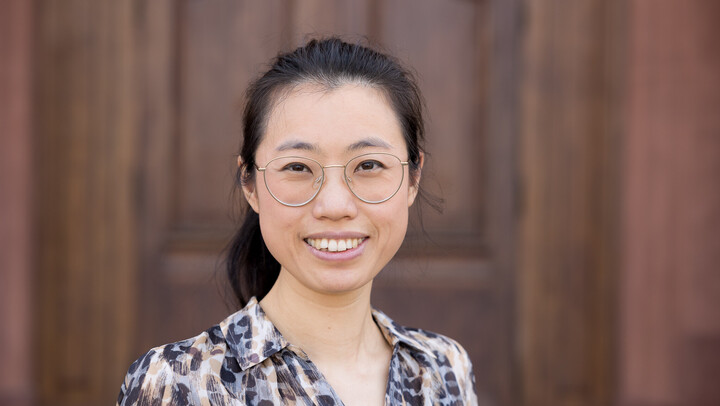 Yufang Sun. She is wearing a white and black blouse and is standing in front of a brown door.