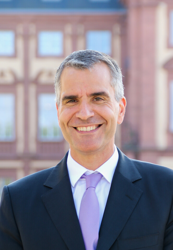 Eine Person trägt ein weißes Hemd, eine lila gemusterte Krawatte sowie einen dunkelblauen Blazer. Die Person heißt Thomas Fetzer.