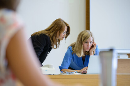 Zwei Frauen schauen zusammen auf den Display eines Laptops. Sie stehen vorne an der Tafel.