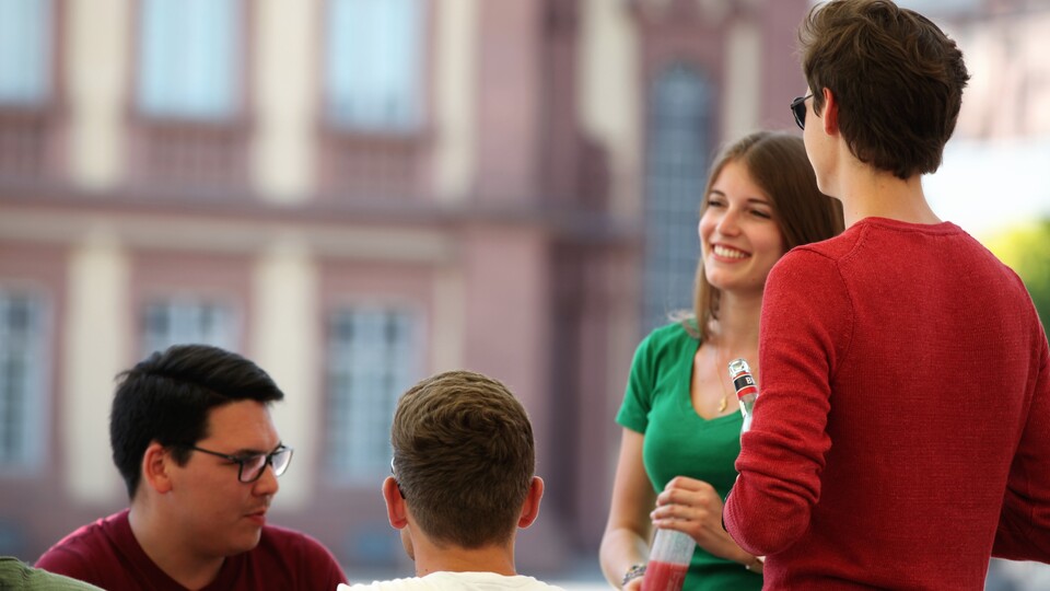 4 Studierende vor dem Schloss. Sie lächeln. Sie unterhalten sich.