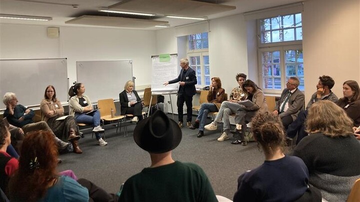 Viele personen sitzen in einem Stuhlkreis. Prof. Dr. Stefan Münzer steht an einem Ende des Kreises und notiert Erkenntnisse auf einem Flipchart.