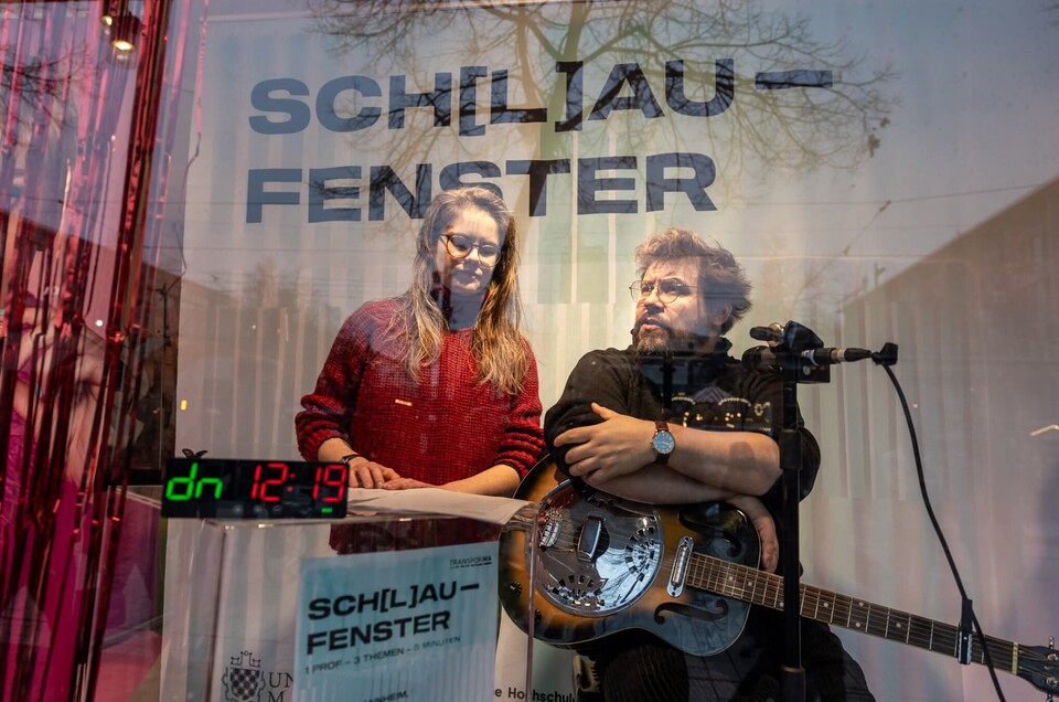 Eine stehende Frau mit langen Haaren und Brille und ein sitzender Mann mit Gitarre und Mikrofon in einem Schaufenster