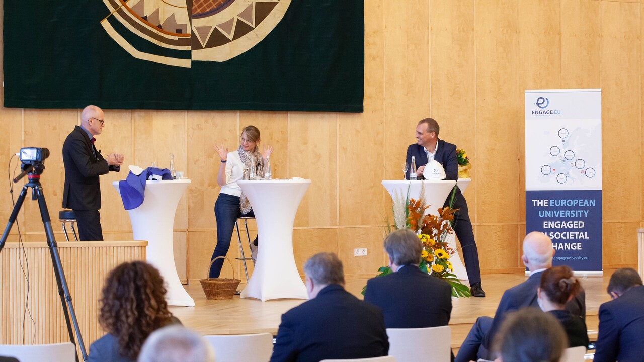 Drei Personen in schicker Kleidung sitzen hinter jeweils einem Stehtisch und unterhalten sich. Im Vordergrund sitzt ein Publikum.