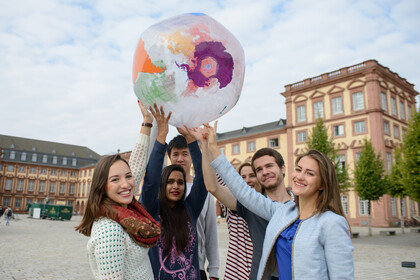 Eine Gruppe Studierender hält eine Weltkugel in die Luft. Sie stehen auf dem Gelände der Unversität Mannheim und lächeln in die Kamera.