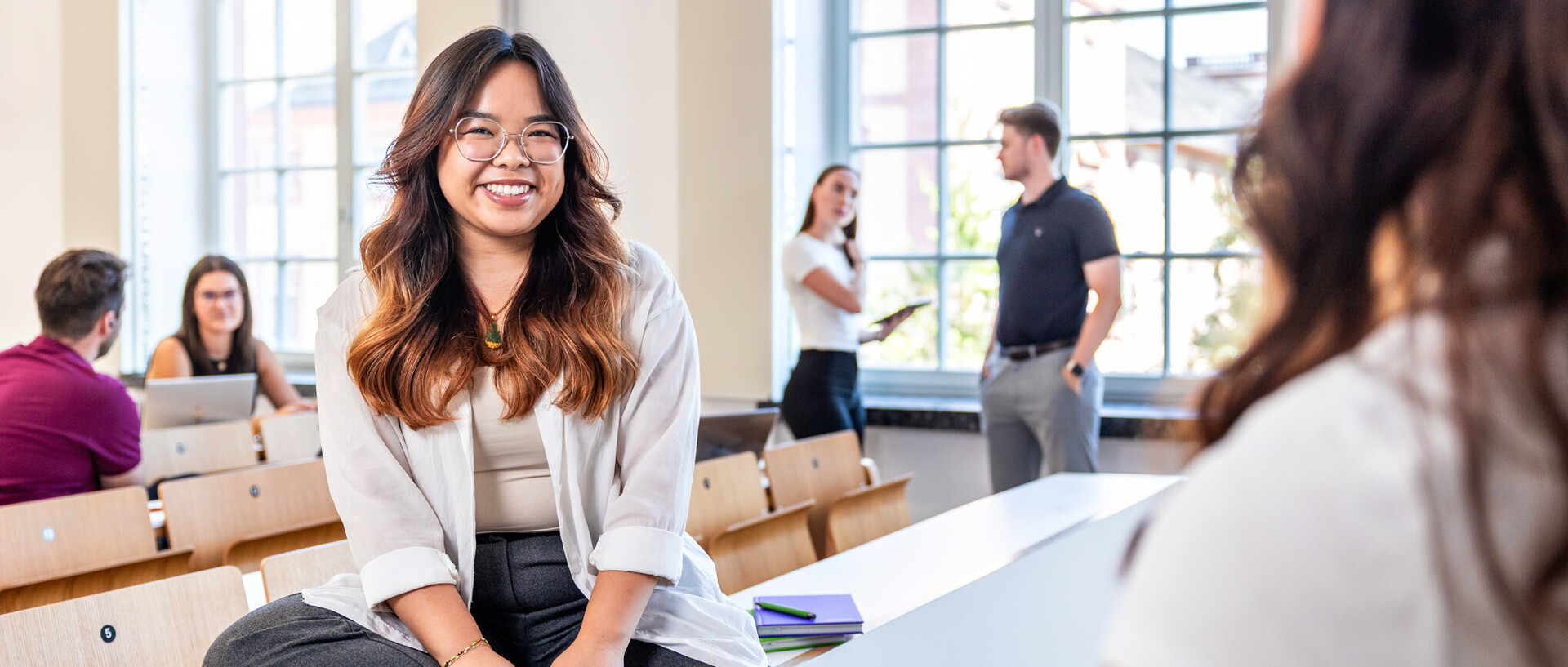 Junge Frau mit braunen welligen Haaren und brille sitzt in einem Hörsaal und lächelt in die Kamera.