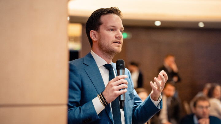 Philipp Bremer trägt einen Anzug, hält ein Mikrofon in der Hand und spricht zu einem Publikum.