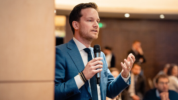 Philipp Bremer trägt einen Anzug, hält ein Mikrofon in der Hand und spricht zu einem Publikum.