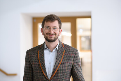 Portrait von Philipp Müller. Er trägt ein weißes Hemd sowie einen braun orange karrierten Blazer und steht in einem gebäude vor einer Glastüre.