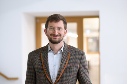 Philipp Müller trägt einen grauen Tweed-Sakko und Brille und lächelt in die Kamera