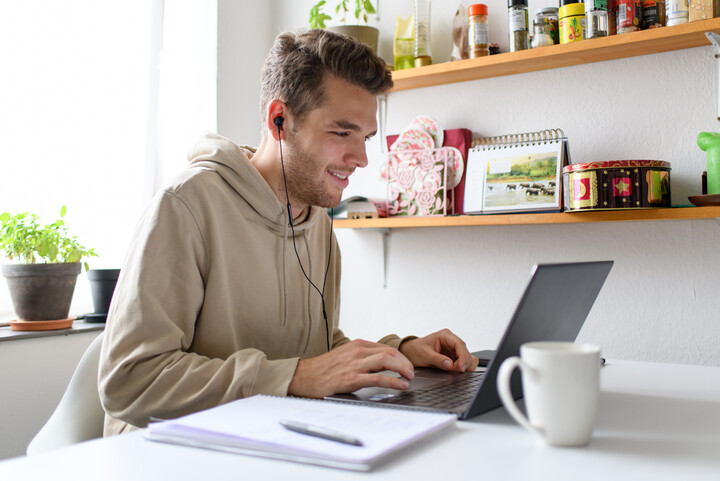 Student in hellbraunen Kapuzenpulli sitzt in seiner Wohnung vor dem Laptop und hat Kopfhörer an. Auf dem Tisch steht eine Tasse und liegt ein Notizblock. Link: Promotion