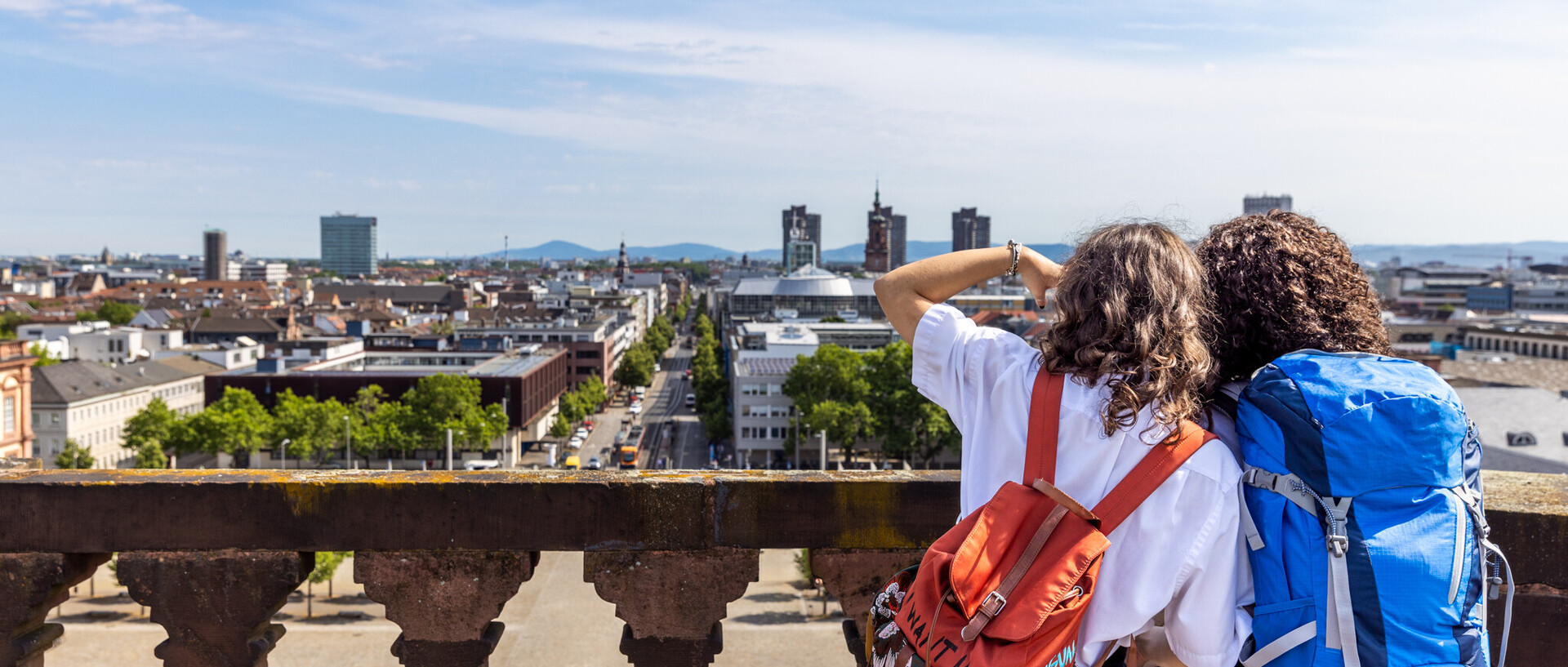 Zwei Studierende mit Rucksäcken auf dem Schlossdach Mannheims. Sie haben einen Blick auf den Ehrenhof und die Stadt.