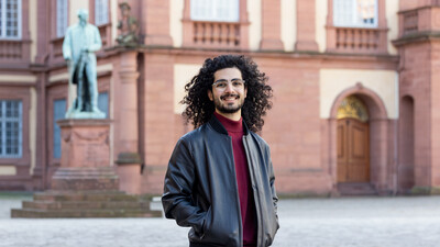 Eliya Ameri hat offene lockige schwarze Haare und lächelt vor dem Schloss in die Kamera. Er trägt eine Brille und eine Lederjacke.