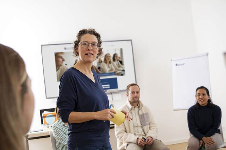 Eine Referentin steht in einem Semiarraum. Um sie herum sitzen erwachsene Personen auf Stühlen und hören zu.