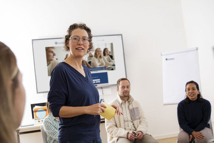 Eine Referentin steht in einem Semiarraum. Um sie herum sitzen erwachsene Personen auf Stühlen und hören zu.